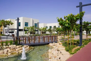 Modern residential community in Sharjah, UAE with villas, landscaped water feature, and pedestrian bridge.
